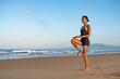 © sutulastock - Fit woman in a black sports outfit performing a balance exercise on sandy beach at sunrise, focused and poised, with soft ocean waves and distant mountains in the background.