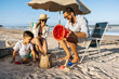 © kleberpicui - A happy family enjoys a sunny beach day as a father helps his son build a sandcastle while the mother relaxes under an umbrella, capturing playful summer moments and seaside bonding by the ocean.