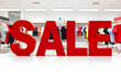 © Pixel-Shot - Red three dimensional sale sign on a white surface in a clothing store with Blurred racks of apparel in the background, Blurred racks of colorful apparel are visible in the background