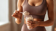 © Treceer - A close-up photograph of a fit woman in brown sportswear preparing a protein shake, holding a shaker bottle and using a scoop to add powder.