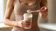 © Treceer - A close-up photograph of a fit woman in sportswear preparing a protein shake, holding a shaker bottle and using a scoop to add powder.