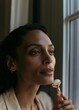 © CYBERPINK - Close-up portrait of a young woman looking out of a window. she is holding a makeup brush in her right hand and appears to be applying foundation or concealer to her face.