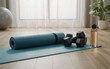 © Shakila - Blue rolled yoga mat with black dumbbells and clear water bottle placed neatly on wooden floor inside bright home gym studio setting during morning sunlight for daily healthy fitness training.