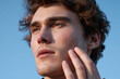 © JoeyMarut - Young man applying moisturizer to jawline with gentle touch in soft natural light, close up portrait showing clear skin texture and contemplative expression against blue sky
