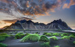 © Travel 'n' Lifestyle - View of Vestrahorn mountain rising above black sand dunes with green grass mounds under a dramatic sunset sky in Hornafjordur, Iceland.