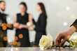 © Pixel-Shot - Hand placing a white rose on a table at a funeral service with blurred grieving people in the background, remembrance and loss