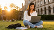 © PrabuanaAndika - Student Using Laptop on University Campus Lawn at Sunset