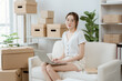 © Wicitr - Entrepreneur's Haven: A young Asian woman, focused and determined, works on her laptop on a comfortable sofa, surrounded by an organized collection of cardboard boxes.