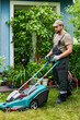 © pressmaster - Vertical image of Caucasian young adult male gardener pushing lawn mower in backyard. Professional landscaping service concept suitable for home maintenance advertising