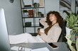 © AnnaStills - Young adult Biracial woman sitting at office desk with legs up holding coffee cup, relaxing during work break. Image suitable for remote work lifestyle and corporate wellness campaigns