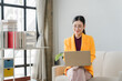 © Kritdanai - Focused Entrepreneur: A confident woman wearing an orange blazer and glasses works diligently on her laptop while seated on a couch in a well-lit living room.