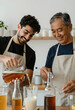 © Pixel-Shot - Smiling young man pouring kombucha into a glass while an older man prepares bottles in a bright kitchen