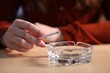 © New Africa - Woman flicking ash from cigarette into ashtray at wooden table indoors, closeup