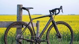 A sturdy mountain bike stands against a wooden post, muddy from an adventurous ride. Bright yellow flowers sway in the gentle breeze under the calm evening sky.