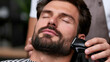 © saichon - Bearded man with closed eyes enjoying professional beard trim by skilled barber using electric trimmer in modern barbershop