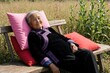 © Viktor - Smiling elderly Asian woman rests on wooden bench in garden. Happy senior lady relaxes with pink, red pillows outdoors. Retired female enjoys peaceful leisure time in nature near farm field during