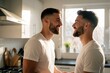 © Maryna - Two young men smile talking in a sunlit kitchen. Casual morning togetherness. They wear white tees, possibly preparing breakfast or coffee at home.