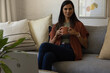 © wavebreak3 - Woman sitting on gray sofa at home holding terracotta mug near laptop and plant, wearing smartwatch