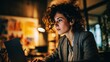 © Harry - Focused young woman with curly brown hair works on her laptop computer wearing a professional grey blazer in dim lighting