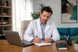 © Studio - Professional doctor in white lab coat writing medical notes on clipboard beside laptop in bright modern office with natural light