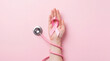 © faysal - A person holding a pink breast cancer awareness ribbon in an open hand with a medical stethoscope wrapped around the arm, set against a solid pink background.