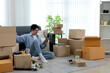 © sabthai - Young Asian man relaxing with tablet while unpacking cardboard boxes in bright new apartment