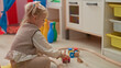 © Krakenimages.com - Girl sits cross legged playing with wooden train track and colorful blocks, hand placing a small car on the track inside a playroom building; concentration curiosity.