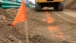 © MSTMAYA - A bright orange marker flag on a construction site with a yellow heavy vehicle in the background, and orange paint, lines on the, dusty ground