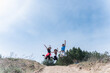 © o_lypa - Two women and a man jump together on a sandy hill during an outdoor hiking trip, expressing joy, energy and friendship against a wide blue sky.