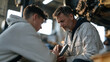 © forenna - Veteran auto mechanic in a rain slicked garage forecourt teaching a teenage apprentice to read a diagnostic scanner, both crouched beside a lifted vehicle, puddles reflecting
