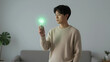 © Lubov - Asian man in a beige sweater holding a glowing green light bulb in a modern home. He looks at the source representing green energy and sustainable innovation
