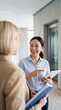 © Pixels Pictures - Smiling Asian businesswoman with tablet explaining project to colleague in modern office hallway