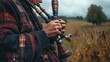 © Halyna - A mature Caucasian male in a plaid jacket plays a woodwind instrument in a field. This image is relevant to Tartan Day