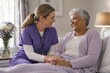 © Lisicus - Female nurse in purple scrubs talking to a smiling senior African American woman lying in bed at home.