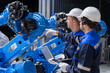© wanakit - Engineers wearing hardhats using laptop and tablet to discuss plans next to robotic arm in factory.