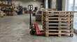 © Bohdan - A sturdy red manual pallet jack stands ready in a vast industrial warehouse supporting a stack of weathered wooden pallets for efficient logistics operations