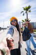 © CarlosBarquero - Vertical. Happy Black woman smiles and reaches forward to lead the viewer by hand, while friends stand behind on a sunny promenade in a modern urban area.