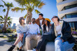 © CarlosBarquero - Diverse friends sit close together in a bright neighborhood and smile during a relaxed outdoor social moment with palm trees and modern architecture.