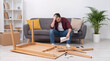 © ChaoticDesignStudio - Frustrated man sitting on sofa while struggling with DIY furniture assembly. Confused homeowner holding manual near broken wooden table in modern living room.