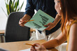 © Studio Marmellata - A business professional holds a green document with bar charts while explaining key data to a colleague in a modern office. They are collaborating on a team project today.