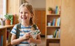 © Nina - Happy 7 year old schoolgirl holding a notebook and pencil smiling in a bright classroom