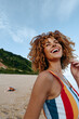 © SHOTPRIME STUDIO - Happy woman with curly hair wearing colorful striped swimsuit smiling outdoors on a sunny beach. Blue sky and green hills in background create a joyful summer mood.