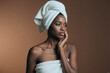 © DragonImages - Young adult Black woman touching cheek after skincare routine, wrapped in towel and presenting self care beauty concept. Ideal for spa, skincare, wellness, cosmetic branding, medium shot