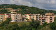 © Guillermo - Vista panorámica de un pueblo mediterráneo con coloridos edificios residenciales en la ladera de una colina verde bajo un cielo nublado.