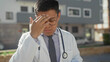 © Krakenimages.com - Man doctor wearing white coat and stethoscope, hand on forehead rubbing temples eyes closed in street near parked cars and building; fatigue concern.