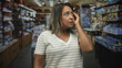 © Krakenimages.com - Woman beckoning with hand while touching temple in store aisle inside building, pensive expression and lifted fingers; concern hesitation.