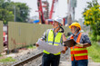 © BESTIMAGE - Two male Construction Workers wearing helmet and vest Discussing Blueprint Plans hold clipboard on a Railway Track Site Du Daylight in a Green Environment with Safety Gear and Tools