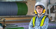 © BESTIMAGE - Confident female worker with earmuffs and safety vest posing with arms crossed in front of weaving machinery in textile factory. Fabric woman supervisor wear uniform working in industrial site.