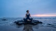 © Setia Fitra - Woman meditating in lotus pose on dark rocks by the ocean at twilight.