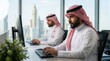 © tanin - Two Saudi men working on computers in traditional attire in a modern office environment